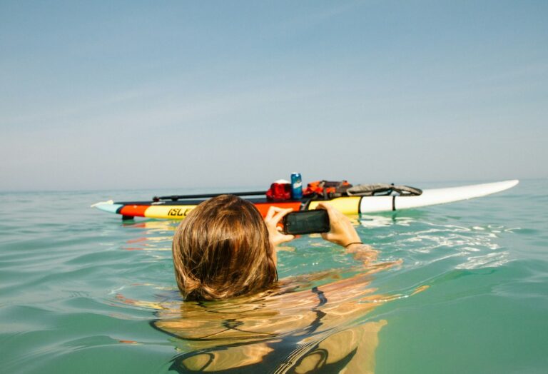 A woman on her phone with her paddle board in the water.